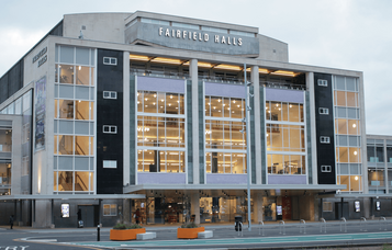 The modern facade of Fairfield Halls in Croydon, showcasing its large glass windows and entrance area under a cloudy sky.